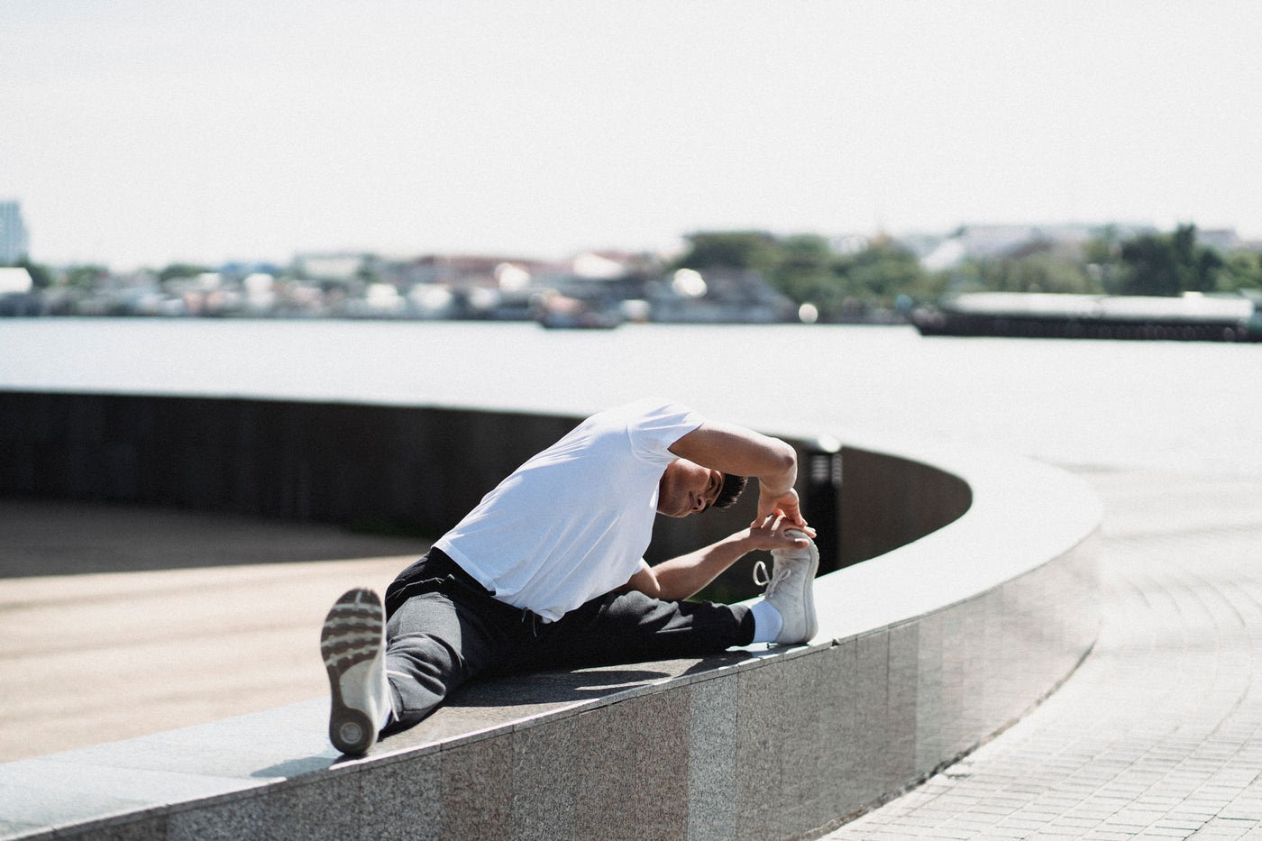Young ethnic male in sportswear stretching legs while sitting on rounded fence near pavement in town