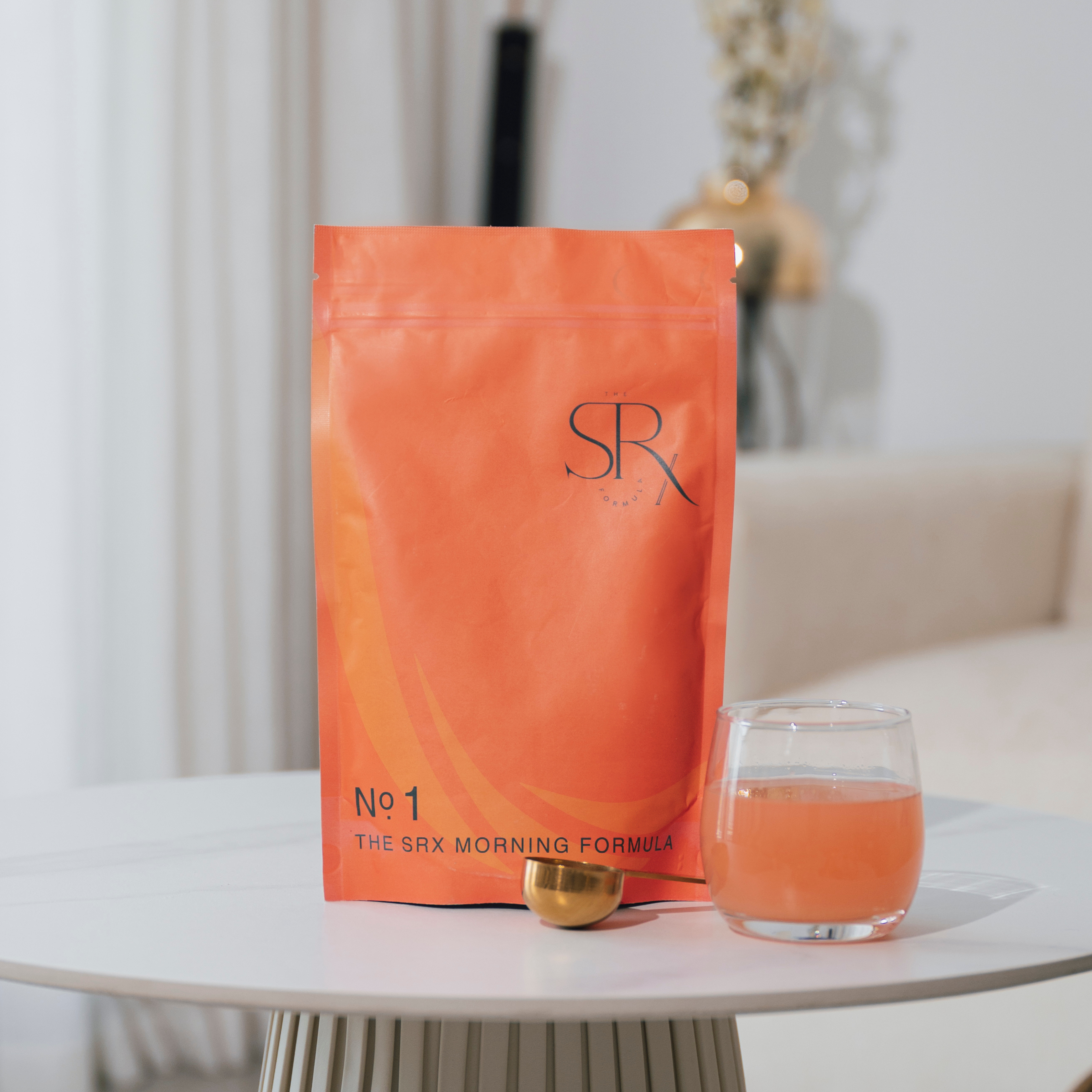 A sealed blue pouch labelled 'SRX' with the text 'No.1 THE SRX MORNING FORMULA' is displayed next to an orange coloured glass of water and a golden object, presented on a white surface with a blurred background.