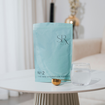 A sealed blue pouch labelled 'SRX' with the text 'No.2 THE SRX HYDRATION FORMULA' is displayed next to a clear glass of water and a golden object, presented on a white surface with a blurred background.